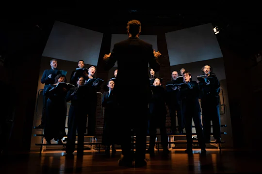 A conductor stands with back to the audience, leading a choir of singers holding sheet music on risers.