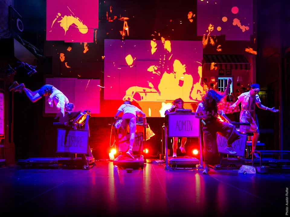Six performers on stage interact with desks labeled "LEISURE," "ADMIN," and "SURVIVAL" against a backdrop of orange and pink digital graphics. Bright lights illuminate the scene.