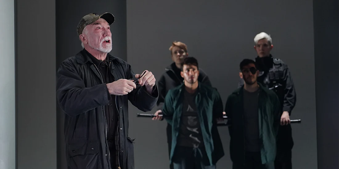 An older man speaks emphatically in the foreground while four people stand in the background, two holding a long rod, all dressed in dark clothing.