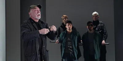 An older man speaks emphatically in the foreground while four people stand in the background, two holding a long rod, all dressed in dark clothing.