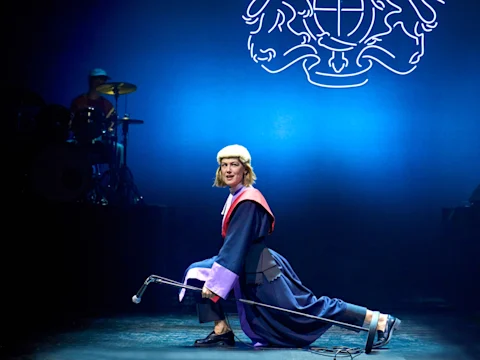 A performer dressed as a judge kneels on stage with a microphone in hand, while a drummer plays in the background under a blue light.