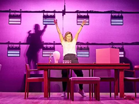 A person stands behind a table with arms raised, surrounded by chairs, drinks, and props, on a stage with bright lighting and a purple backdrop.