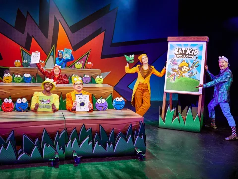 A live stage performance featuring actors dressed in colorful costumes, presenting a scene from "Cat Kid Comic Club." Some hold books while one displays a large board with the show's title.