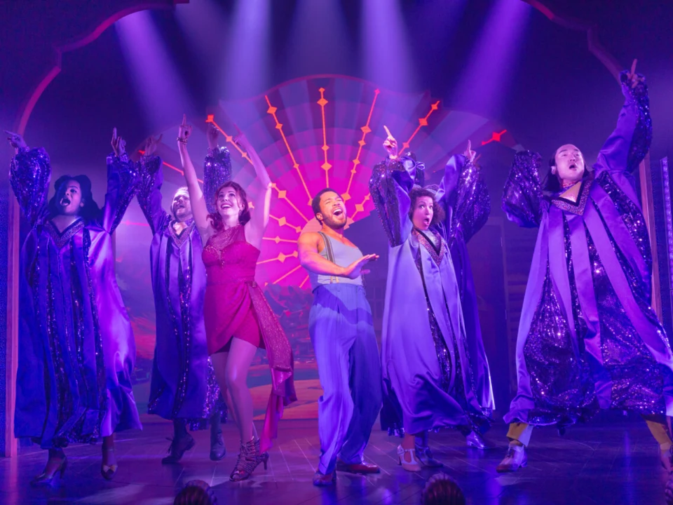 Performers on stage in colorful costumes dance energetically, surrounded by dramatic lighting and a vibrant backdrop.