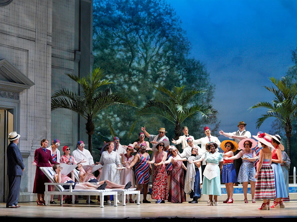 A group of people in colorful period clothing gather on a stage with palm trees and a blue sky backdrop. Some stand, while others sit on lounge chairs, all appearing engaged in conversation.