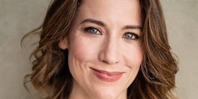 A woman with wavy brown hair and blue eyes smiles softly at the camera against a neutral background.