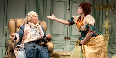 A woman in a period costume gesturing toward an elderly man sitting in a wingback chair.