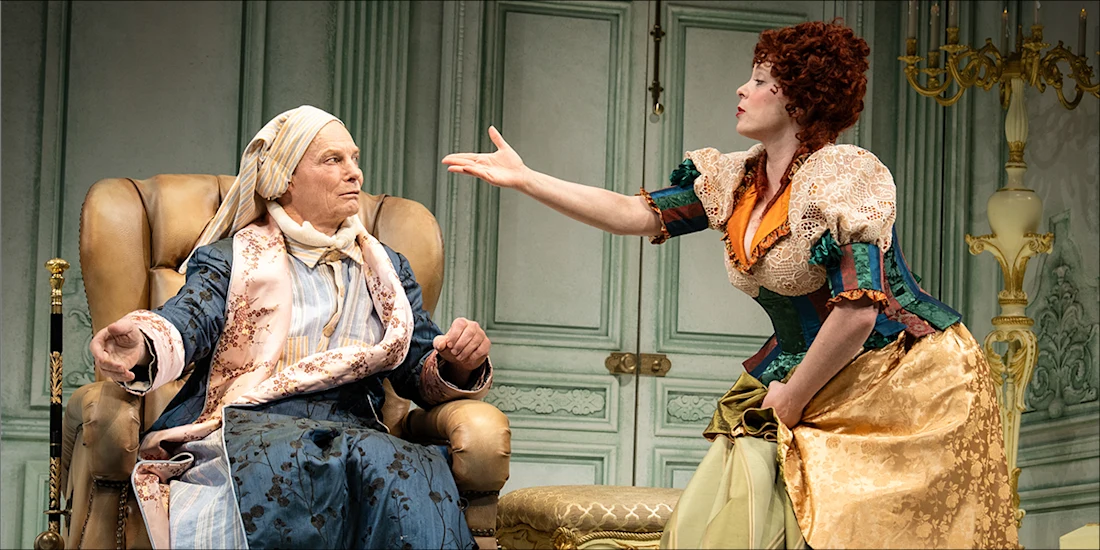 A woman in a period costume gesturing toward an elderly man sitting in a wingback chair.