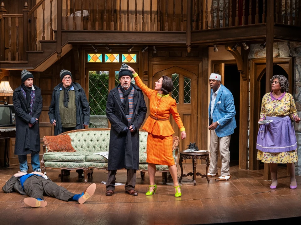 Actors on stage perform a dramatic scene in a living room set. A woman in orange adjusts a man's hat while others watch. One person lies on the floor.