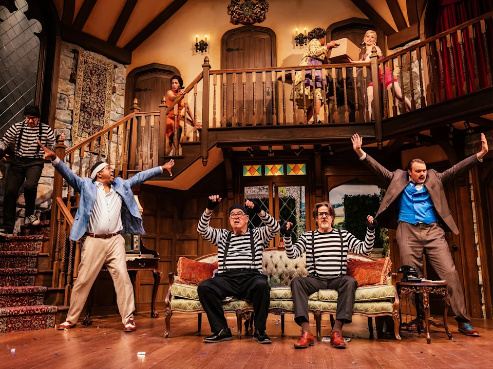 Actors on stage perform a dramatic scene in a living room set. A woman in orange adjusts a man's hat while others watch. One person lies on the floor.