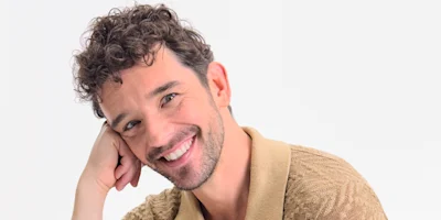 A man with short curly brown hair and facial hair, wearing a beige collared shirt, smiles while resting his head on his hand against a plain white background.