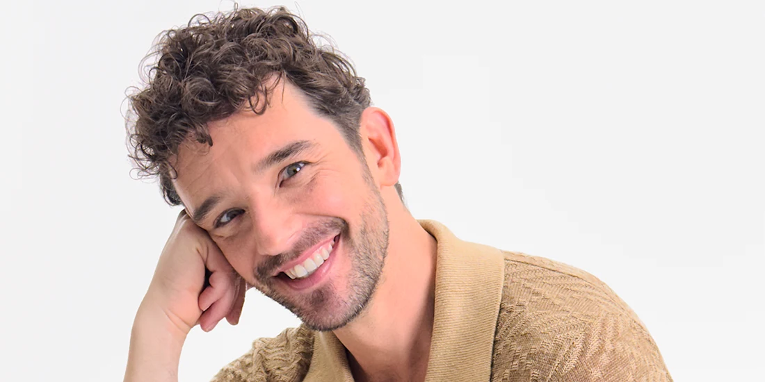 A man with short curly brown hair and facial hair, wearing a beige collared shirt, smiles while resting his head on his hand against a plain white background.
