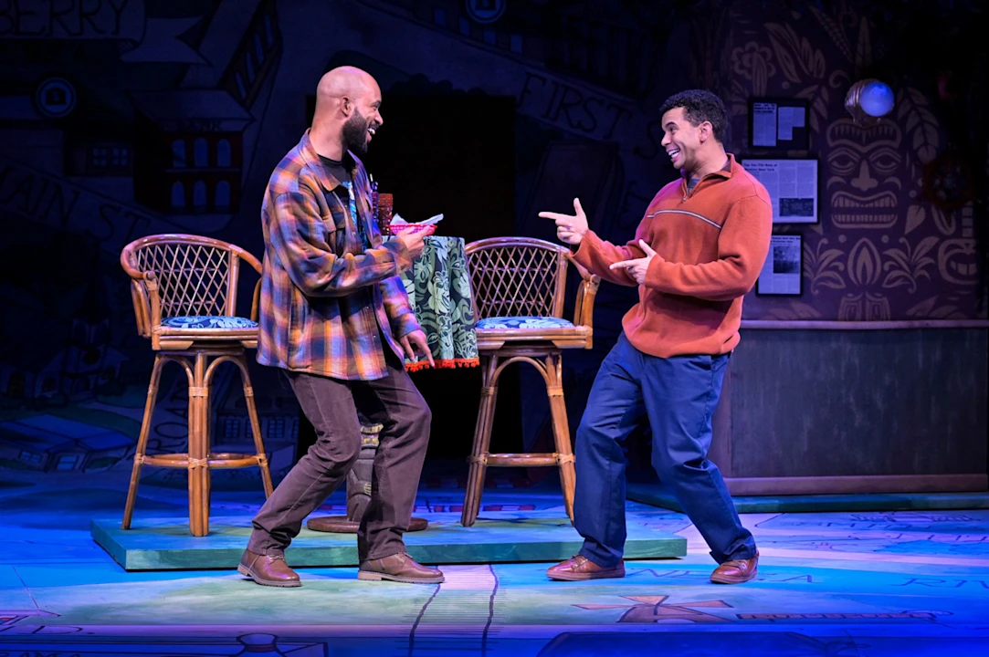 Two men stand on stage, smiling and pointing at each other, with bar stools and a colorful backdrop behind them.