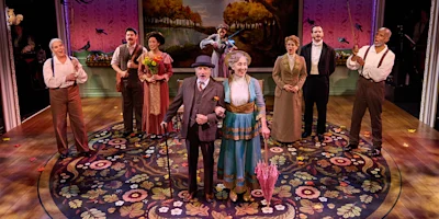 Nine actors in period costumes stand on a stage with a floral carpet and painted backdrop, two in front holding hands, others smiling or clapping in the background.
