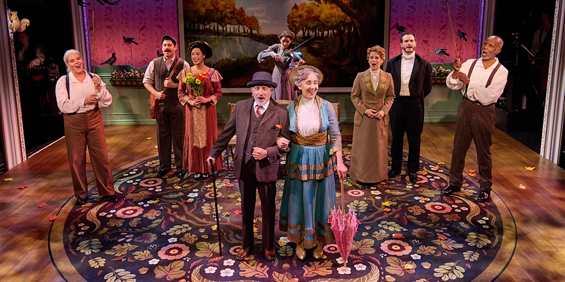 Nine actors in period costumes stand on a stage with a floral carpet and painted backdrop, two in front holding hands, others smiling or clapping in the background.