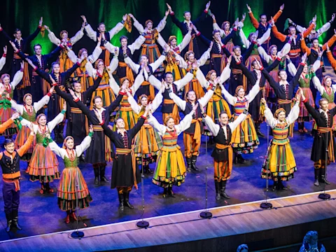 A large group of performers in colorful traditional folk costumes stand on stage with arms raised in a V shape during a choreographed dance.