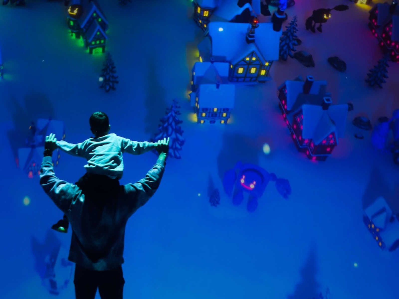 A person holds a child on their shoulders, looking at a miniature winter village display with colorful lights.