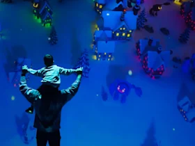 A person holds a child on their shoulders, looking at a miniature winter village display with colorful lights.