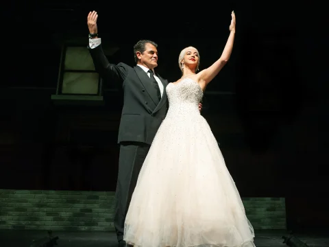 A man in a black suit and a woman in a white gown stand on stage, holding one arm up each.