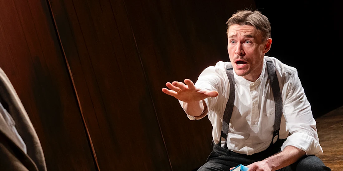 A man in suspenders and a white shirt sits indoors, extending his right arm forward with an expressive, intense facial expression.