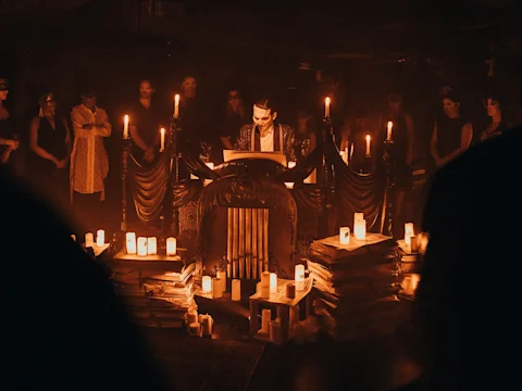 A person stands at a draped podium surrounded by candles and stacks of papers, with a group of people observing in a dimly lit, ritualistic setting.