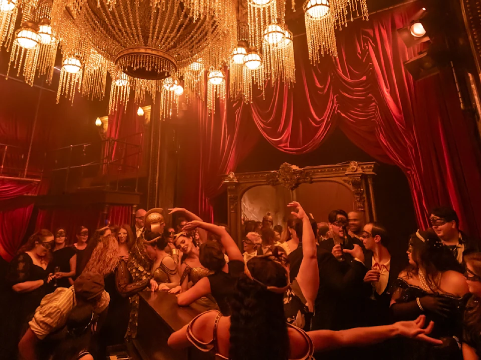 People in formal, masked attire gather under a large chandelier in a dimly lit room with red drapes and ornate decor, creating an atmosphere of a masquerade ball.