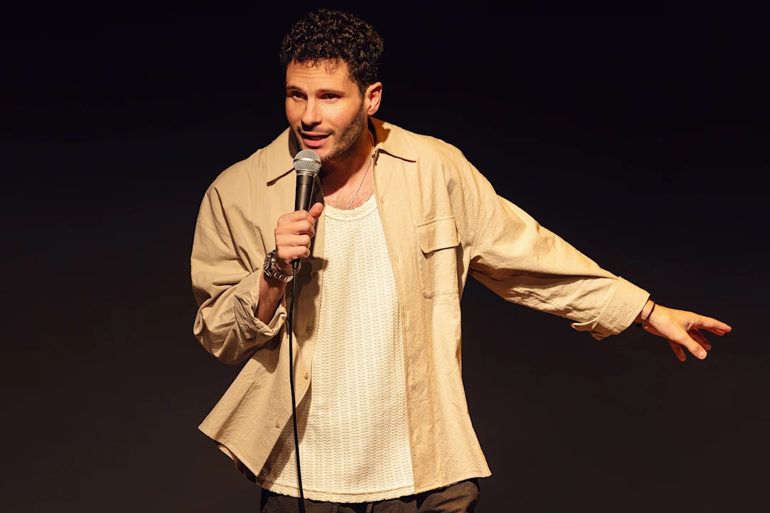 A man performs stand-up comedy on stage, holding a microphone and gesturing to the side.