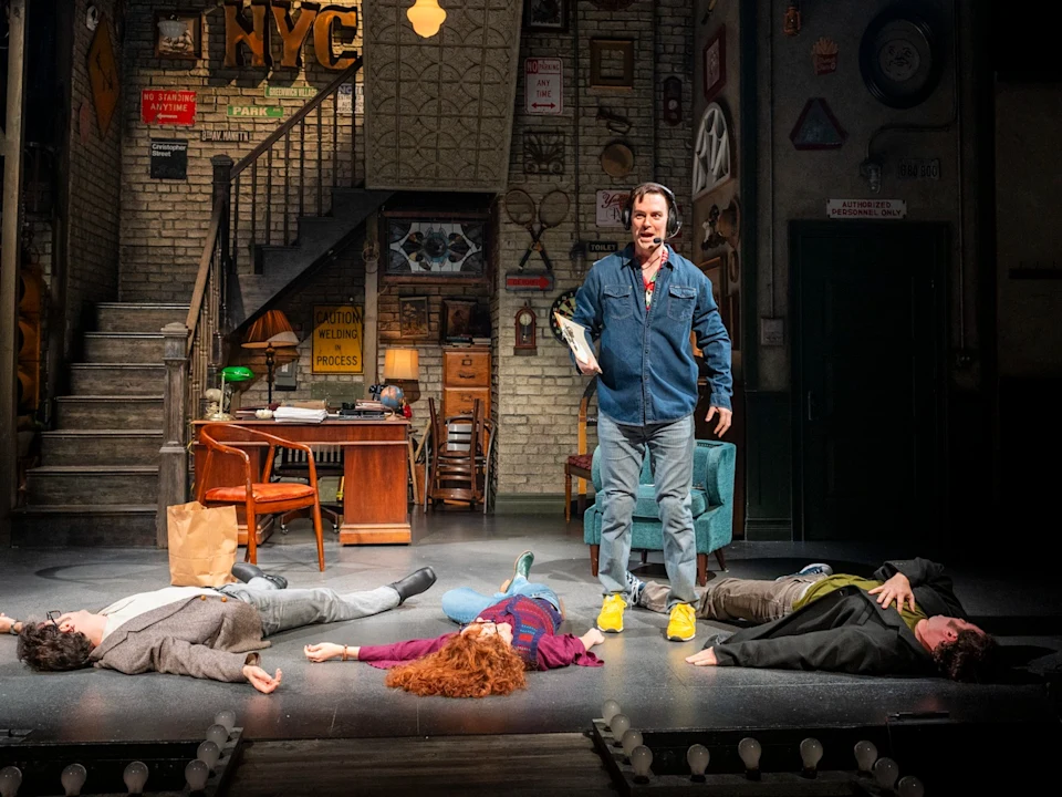 A man stands in an office set holding a folder while three people lie on the floor around him, suggesting a dramatic scene in a stage play.