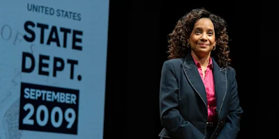 Woman in a dark suit and pink shirt stands on stage next to a sign reading "United States State Dept. September 2009.