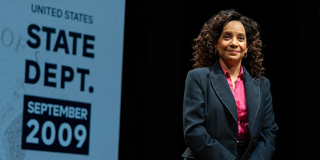 Woman in a dark suit and pink shirt stands on stage next to a sign reading "United States State Dept. September 2009.