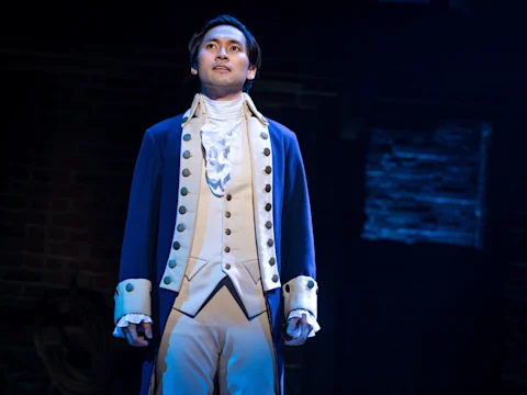 A man in 18th-century style theatrical costume stands on stage under dramatic lighting, gazing upward with a serious expression.