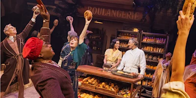 Actors on stage in a bakery setting, joyfully holding up bread, with bakers behind the counter and shelves full of loaves in the background.