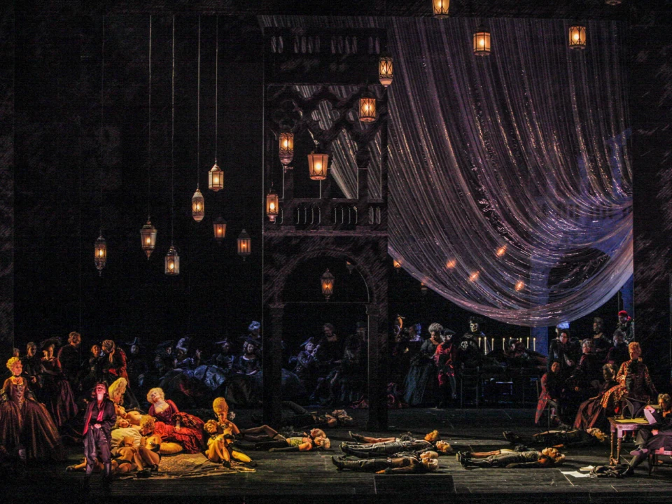 A dimly lit stage features actors in period costumes amidst scattered bodies and hanging lanterns, with an audience in the background and a draped curtain to the right.