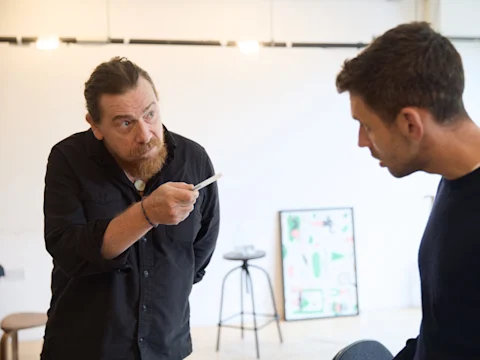 A bearded man gestures animatedly while speaking to another actor during rehearsal.
