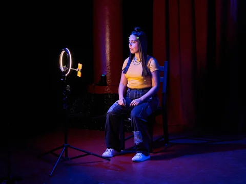 A person sits on a chair under a ring light, wearing a yellow top and jeans, in a dimly lit room with red curtains.