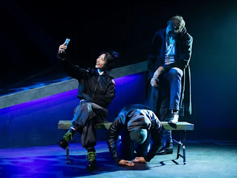 Three people on stage: one sits and takes a selfie, another kneels with head down, and a third sits hunched over at the end of a bench under dramatic lighting.