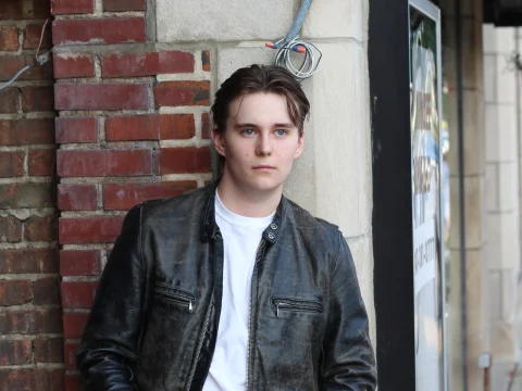 A person with short hair wearing a white shirt and dark leather jacket stands against a brick and cement wall.