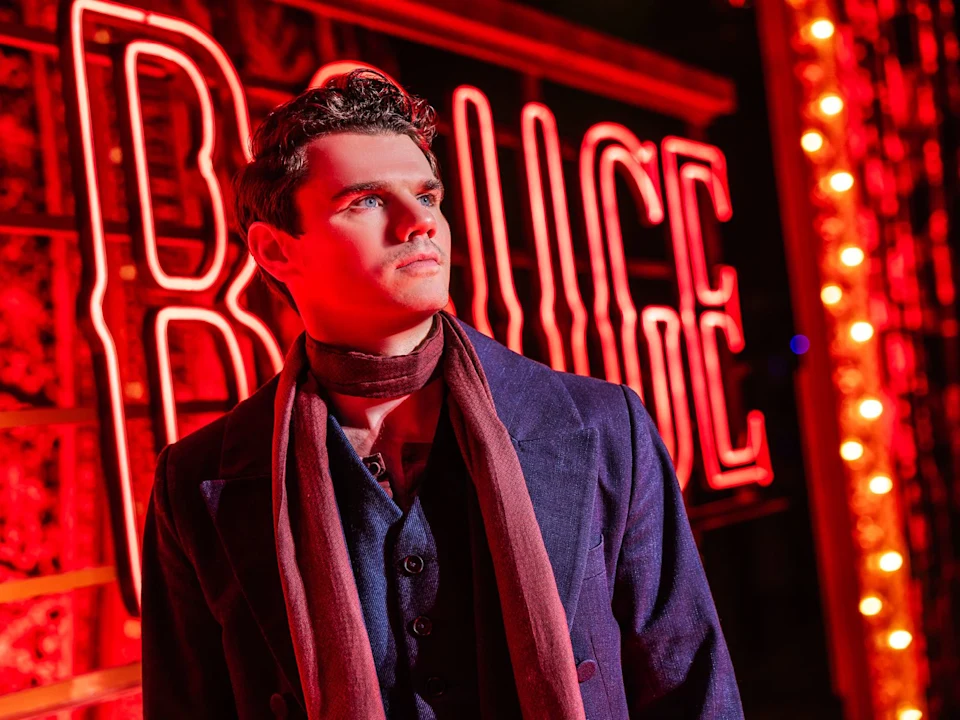A man in a dark suit and scarf stands in front of a brightly lit red neon sign that says "ROUGE.
