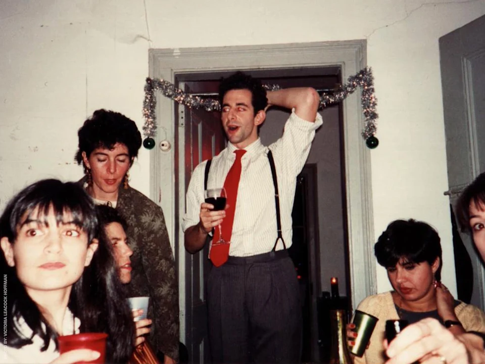 A group of people at a party, some holding drinks. One person in a shirt and tie stands, raising an arm, in a room decorated with tinsel.