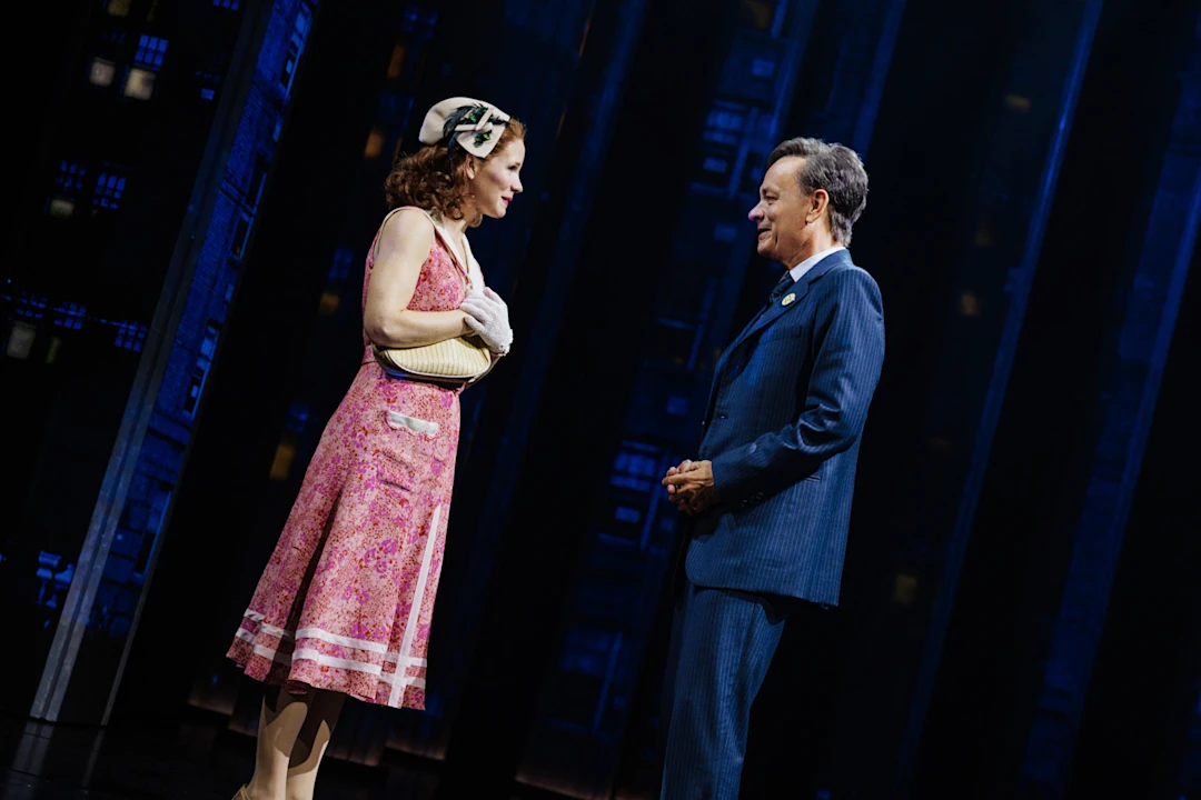 Two actors stand facing each other on stage; the woman wears a pink dress and hat, the man wears a blue suit. The backdrop features dark, tall building facades.