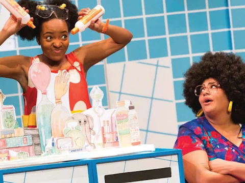 Two actors in a stage production perform behind a counter decorated with cartoon drawings of laboratory items.