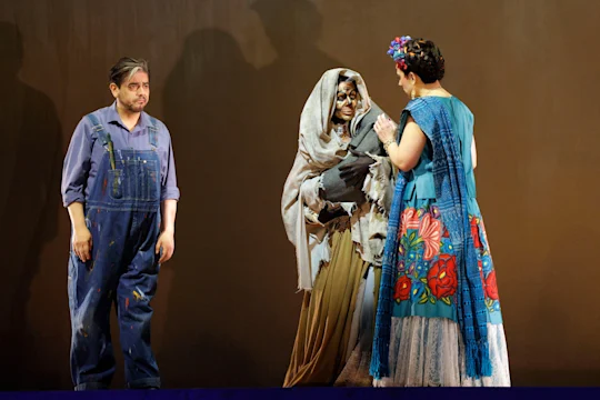Three actors on stage: one in overalls, one in ragged clothing and a hood, and one in a colorful floral dress with a blue shawl, standing against a plain backdrop.