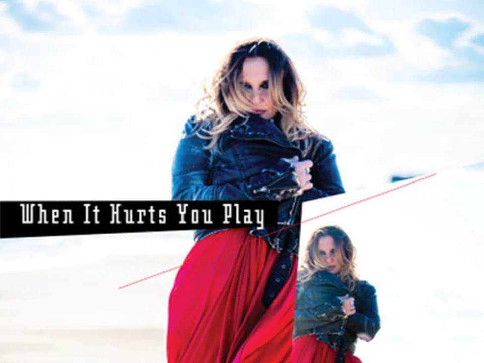 A woman with long hair stands on a beach wearing a black jacket and red skirt.