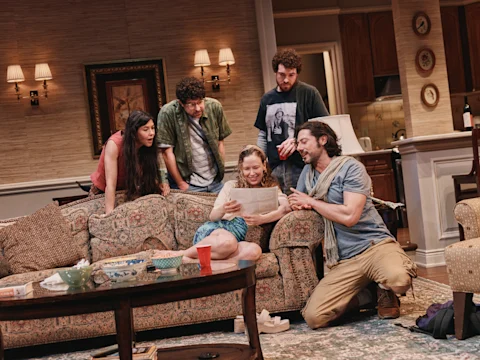 Five people in casual clothes gather around a woman sitting on a couch, looking at a paper together in a cozy, well-lit living room setting.