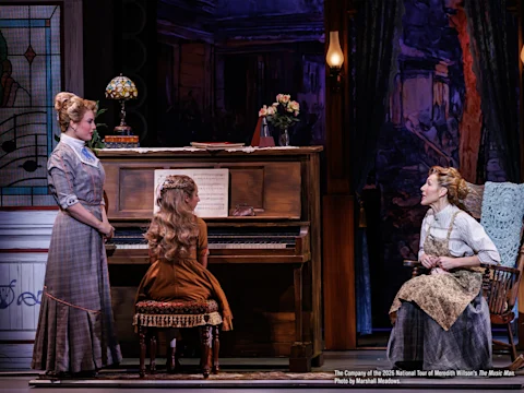 Three women in period costumes are on stage; one plays the piano while the other two stand and sit nearby, engaged in conversation in a theatrical setting.