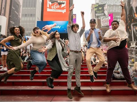 Six people jump in the air with excited expressions on red steps in a busy city area, surrounded by billboards and a crowd.