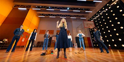 A group of people stand on a gymnasium stage, wearing denim outfits, as one person at the center gestures with a raised finger.