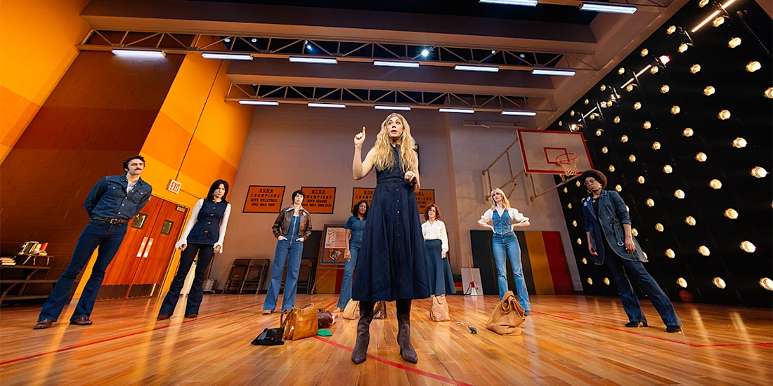 A group of people stand on a gymnasium stage, wearing denim outfits, as one person at the center gestures with a raised finger.