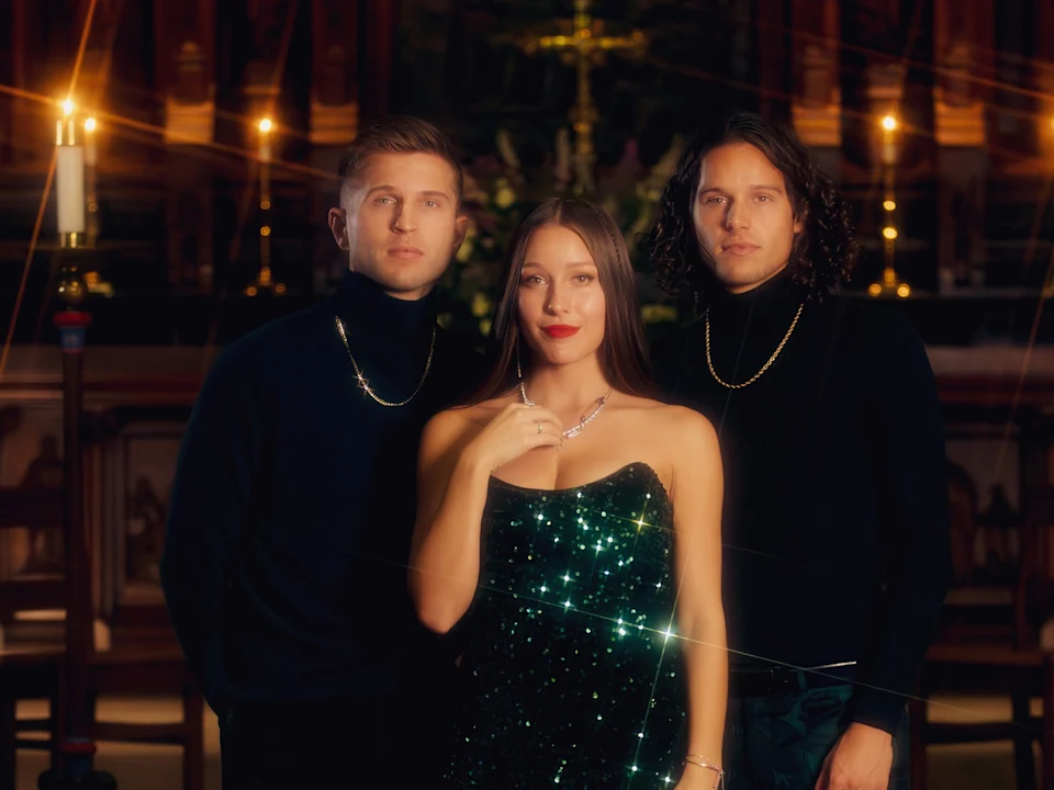 Three people pose indoors; two men in black turtlenecks with gold chains stand beside a woman in a sparkly green dress, with candles and ornate decor in the background.