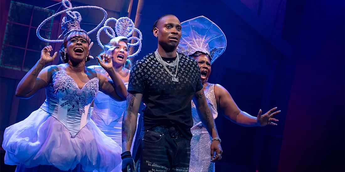 A man in casual clothes stands center stage as three women in elaborate white costumes and headpieces sing behind him under stage lighting.